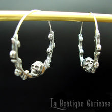 Laden Sie das Bild in den Galerie-Viewer, Boucles d'oreilles gothiques, style créoles, en acier inoxydable. Branche en forme de cercle ornée de petits crânes sur laquelle repose une tête de mort plus grande que les autres. Esprit romantique, memento mori.