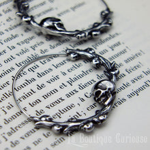 Laden Sie das Bild in den Galerie-Viewer, Boucles d'oreilles gothiques, style créoles, en acier inoxydable. Branche en forme de cercle ornée de petits crânes sur laquelle repose une tête de mort plus grande que les autres. Esprit romantique, memento mori.
