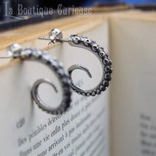 Charger l'image dans la galerie, Paire de boucles d'oreilles tentacules de pieuvre arrondies en acier posée sur un livre ancien.