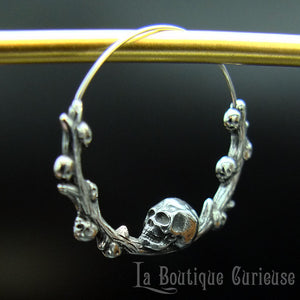 Boucles d'oreilles gothiques, style créoles, en acier inoxydable. Branche en forme de cercle ornée de petits crânes sur laquelle repose une tête de mort plus grande que les autres. Esprit romantique, memento mori.