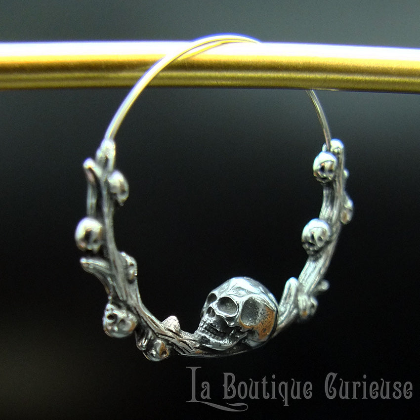 Boucles d'oreilles gothiques, style créoles, en acier inoxydable. Branche en forme de cercle ornée de petits crânes sur laquelle repose une tête de mort plus grande que les autres. Esprit romantique, memento mori.