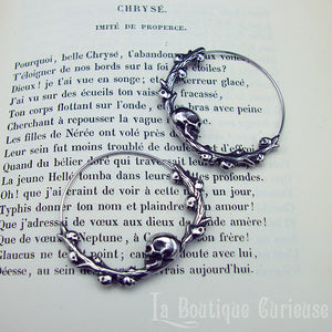 Boucles d'oreilles gothiques, style créoles, en acier inoxydable. Branche en forme de cercle ornée de petits crânes sur laquelle repose une tête de mort plus grande que les autres. Esprit romantique, memento mori.