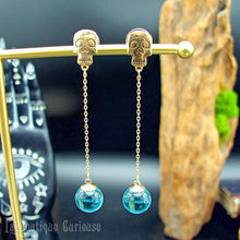 Laden Sie das Bild in den Galerie-Viewer, Paire de boucles d’oreilles pendantes dorées avec crânes et sphères bleues en verre.