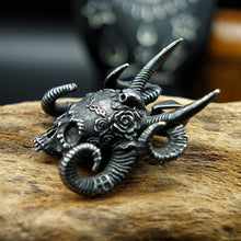 Charger l'image dans la galerie, Pendentif crâne gothique aux cornes de démon en acier inoxydable noir avec cornes et couronne de roses vu de profil posé sur du bois.